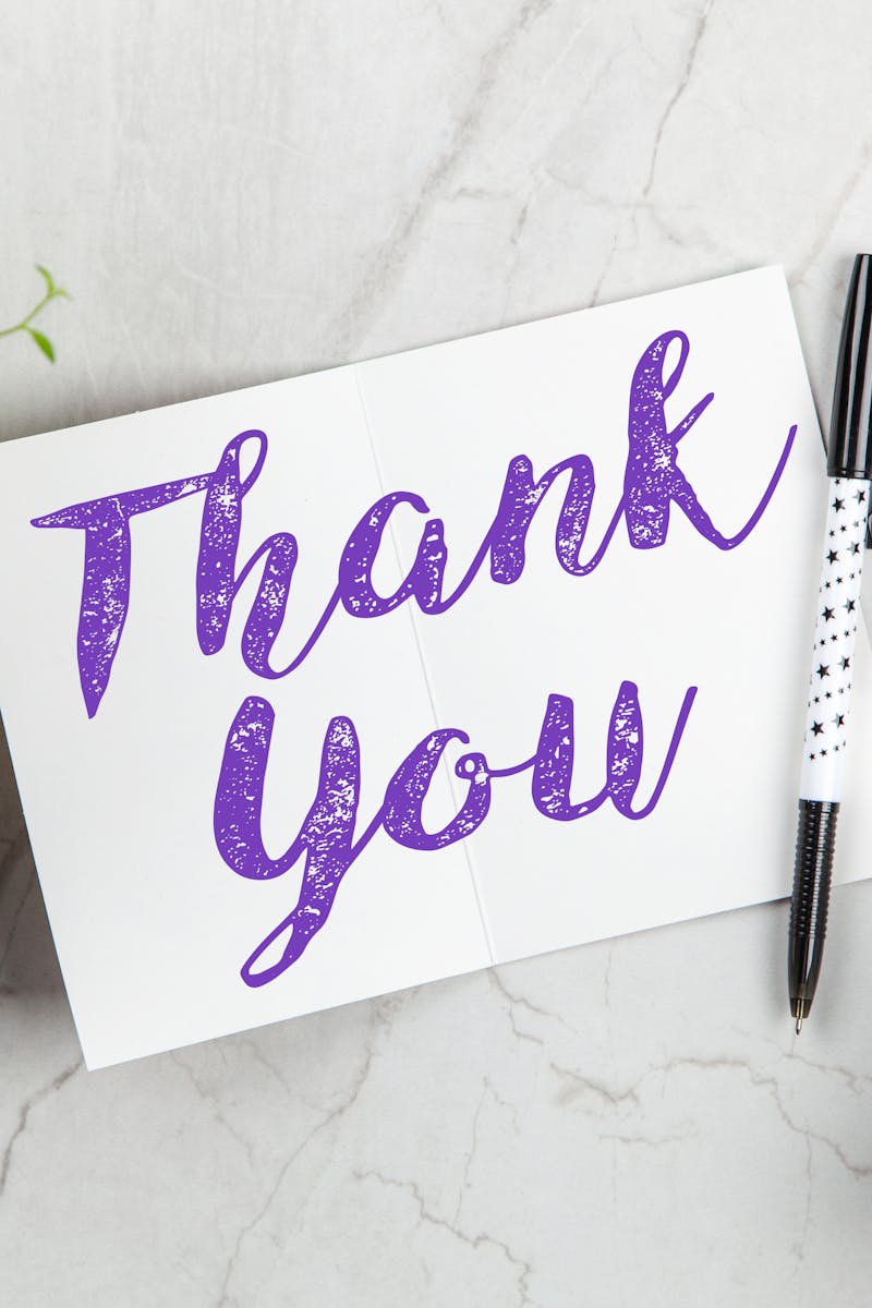 A stylish thank you card paired with purple flowers, a gift box, and pen on a marble surface.